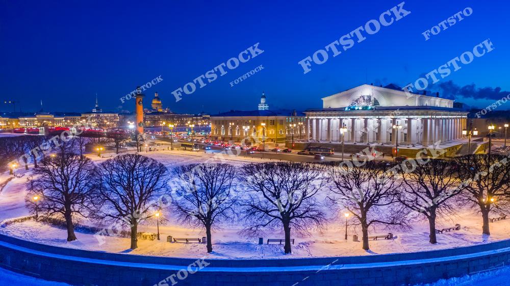 На фотографии изображена Стрелка Васильевского острова в зимний вечер. Ростральные колонны и здание Биржи подсвечены огнями, подчёркивая их величие. Снежный покров придаёт пейзажу особую атмосферу. Вдалеке видны Исаакиевский собор и другие достопримечательности Санкт-Петербурга.